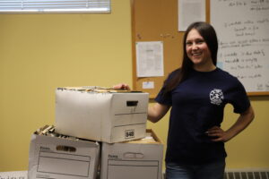 Woman stands next to boxes of old papers