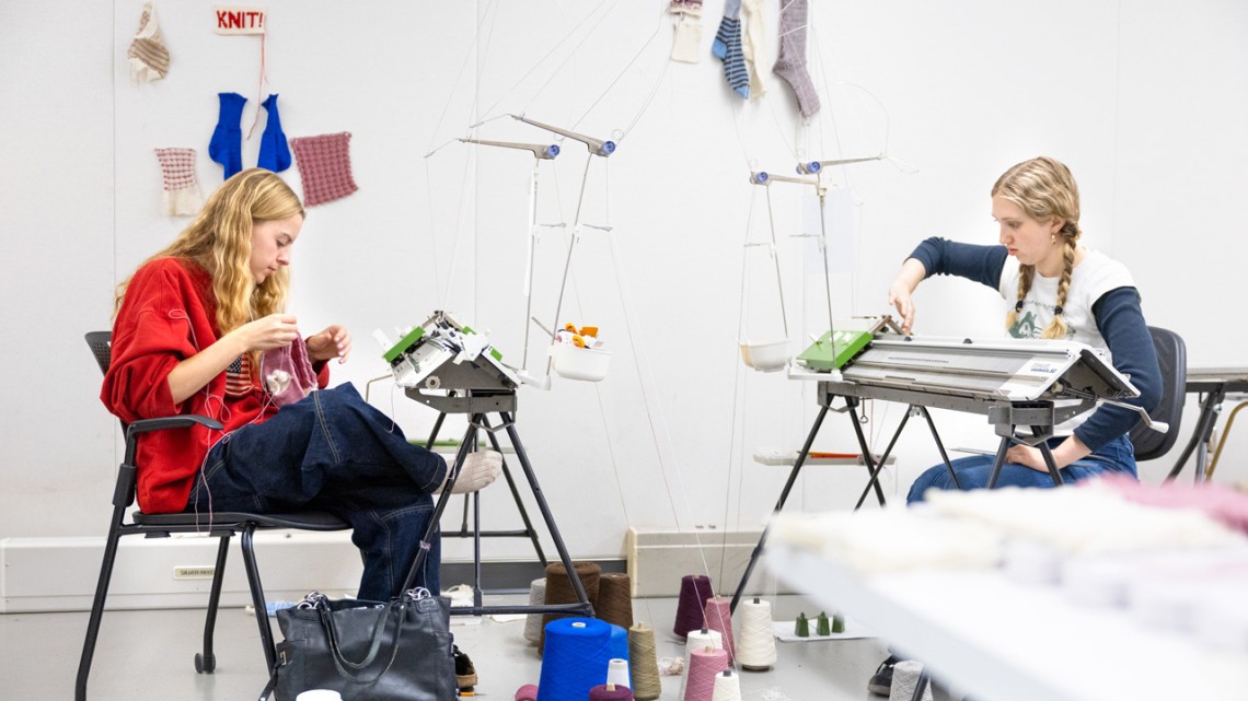 Fashion design majors Roan Harvey ’27, left, and Lucy Jones ’26 make knitted socks using wool grown and spun in New York state for the class Knitwear Design and Other Applications in the College of Human Ecology. Their design swatches and test socks are pinned to the wall. Alexandra Bayer/Cornell University