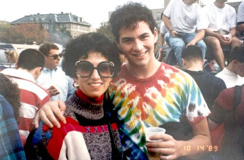 Portrait of two people, mother and son, in the middle of a crowd. Mother, left, smiles at camera with large sunglasses. Son, right, smiles at camera in rainbow tie dye shirt